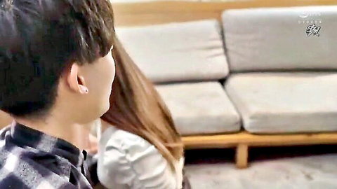East Asian man in checkered shirt cuddles long-haired woman in white blouse on gray couch