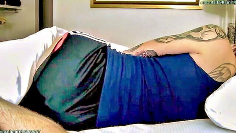 Tattooed muscular guy lying on bed in tight shiny blue shorts and tank top