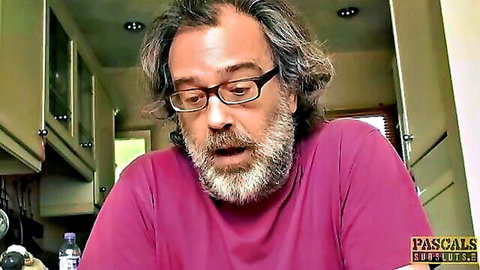 Mature bearded man with glasses in pink turtleneck sits in kitchen