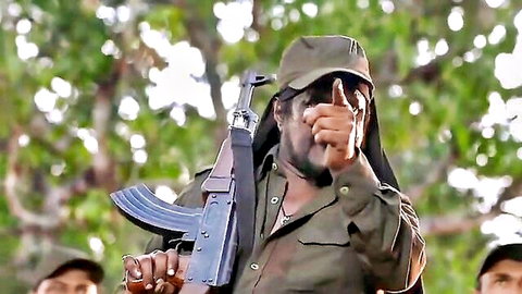 Armed militant in green uniform shushing with finger to lips holding AK-47 outdoors