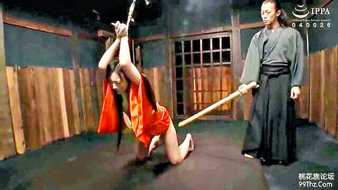 Japanese woman in red kimono bound shibari-style kneels submissively arms overhead as man in robes holds staff BDSM domination