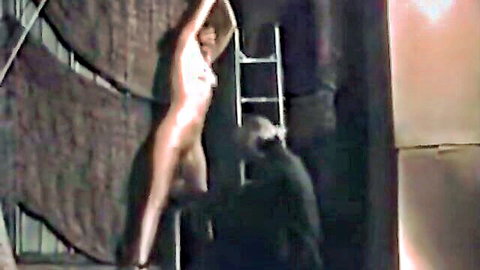 Naked slim woman suspended arms up in BDSM scene with man nearby