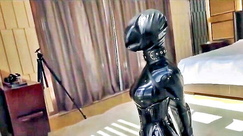 Woman in shiny black latex catsuit, hood, and collar posing in BDSM fetish scene