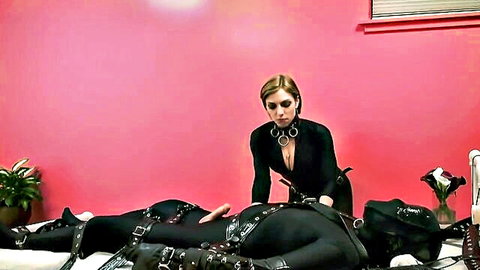 Blonde femdom in latex catsuit teases bound male sub in BDSM domination