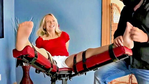 Smiling blonde MILF strapped legs spread wide on red BDSM bondage bench in shorts