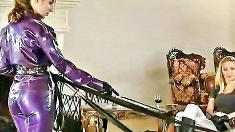 Femdom mistress in shiny purple latex leashes collared blonde submissive in BDSM dungeon