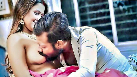 Topless Indian MILF smiles as bearded man sucks her big tits nipple on bed