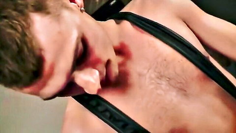 Brunette guy with red marks on face and chest wearing black leather straps in rough BDSM scene