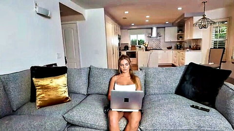 Blonde babysitter in white tank top sitting legs spread on couch with laptop