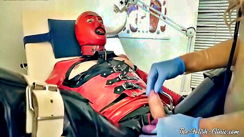 Gloved femdom hand strokes hard cock of red latex bound sub on fetish clinic exam table