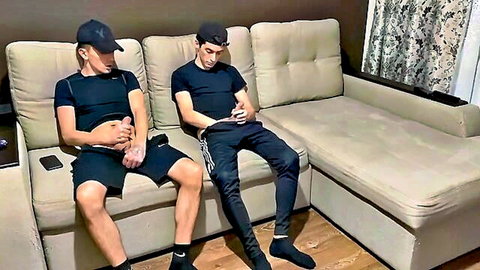 Two young twinks lounging on couch one lifting shirt exposing abs gripping crotch bulge