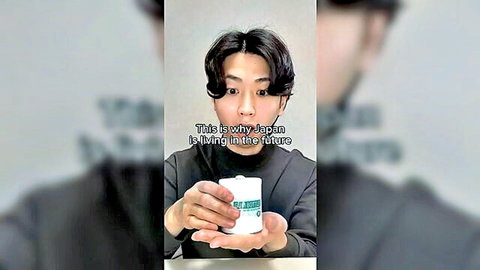 Young Asian man in gray shirt holding white mug with green logo, surprised expression