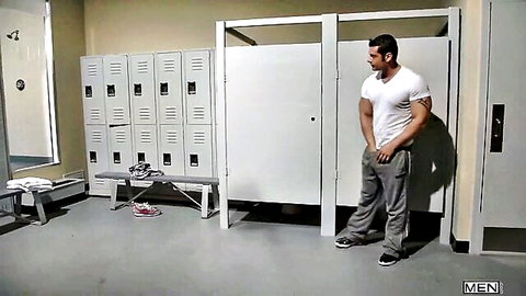 Muscular hunk in white tee and gray sweatpants stands in gym locker room near showers