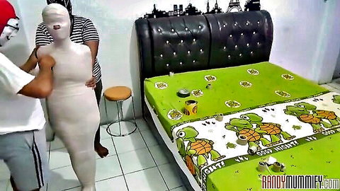 Two men handling tightly mummified bound woman in BDSM bondage threesome on green turtle bed