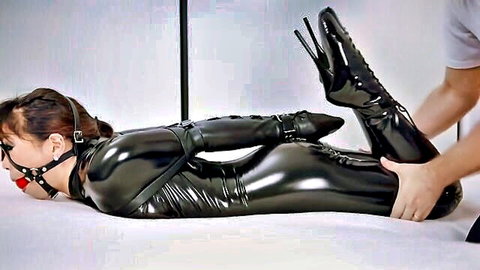 Bound Asian woman in shiny black latex catsuit gagged with ball gag as man grips her stiletto heels in intense BDSM bondage