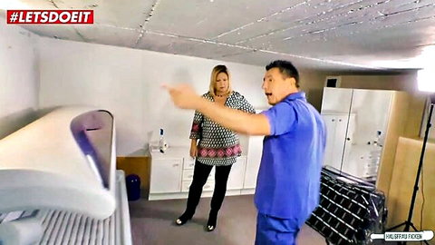Excited mature blonde woman in tight pants and heels gestures with smiling Asian man in blue scrubs beside tanning bed