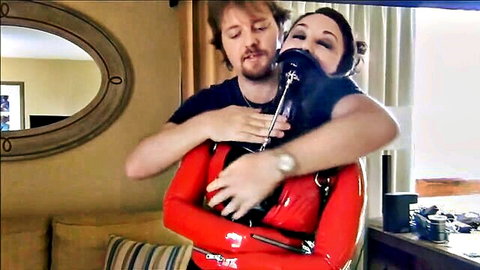 Brunette MILF in tight red latex catsuit with ball gag hugged from behind by bearded man