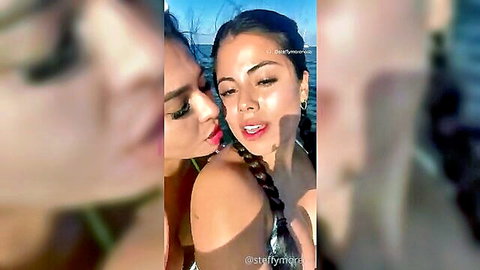 Two tanned Latina brunettes in bikinis kiss cheeks passionately outdoors by the water