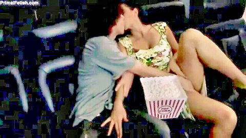 Busty woman in floral dress straddles man kissing passionately in dark theater holding popcorn bucket on lap