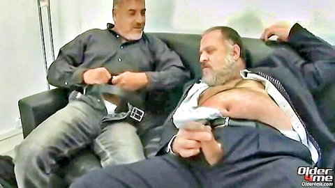 Two older bulky men in intimate pose, one with open shirt exposing hairy chest, other unbuckling jeans
