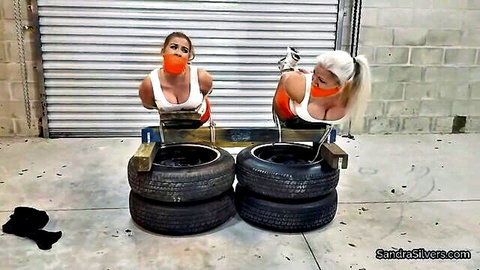 Two gagged busty blondes and brunettes in bondage chairs surrounded by tires teasing underboobs