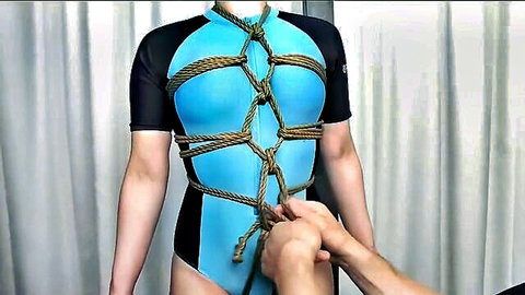 Asian woman in blue bodysuit bound in intricate shibari ropes, man tying knots