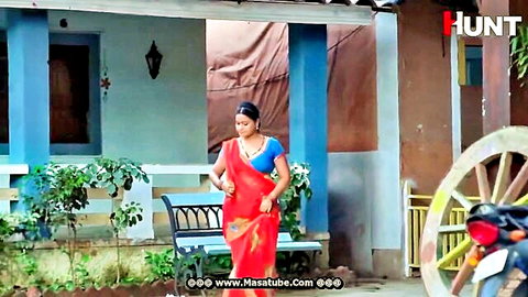 South Asian woman in red saree standing outdoors by blue house with plants and bench