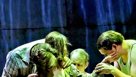 Four green-haired men huddled intimately touching faces on dark stage with blue curtain