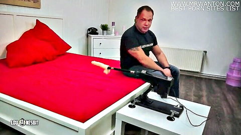 Bald man in black shirt sits by red bed with fucking machine and thick white dildo attached