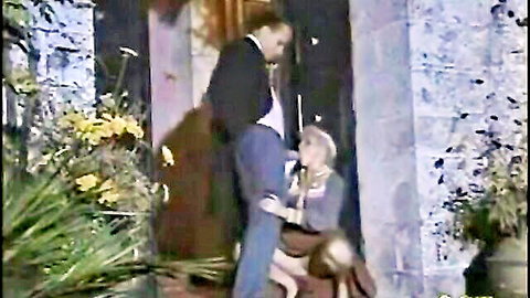 Blonde teen kneels sucking older man's cock outdoors by flowery door.