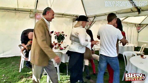 Curvy blonde BBW MILF in hat kissing hunk at outdoor tent party with suited men