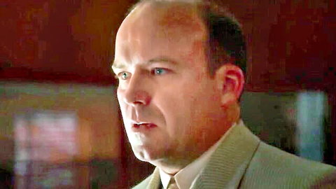 Balding man in light suit staring intensely with open mouth in dim room