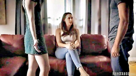 Brunette in short green dress stands with seated woman in jeans and man, teasing threesome vibe