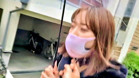 Cute Asian woman with brunette hair in pink mask holding umbrella outdoors teasingly