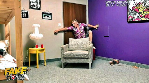 Blonde guy in pink Hawaiian shirt jumps Superman pose over armchair in colorful bedroom