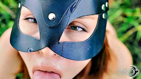 Redhead in black leather cat mask teases with tongue out outdoors