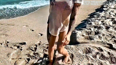 Michaela Isizzu in sheer beach dress, tattoos, standing on sandy shore by ocean