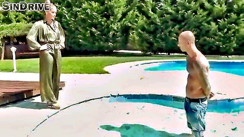 Blonde in shiny green jumpsuit teases shirtless tattooed bald hunk by the pool