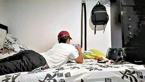 Young Asian guy in red cap lying sideways on bed holding phone in amateur bedroom