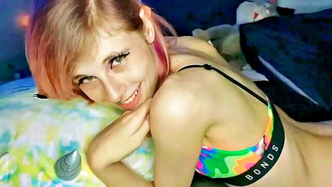 Smiling pink-haired woman in colorful bra on bed, soft tease pose
