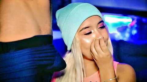 Blonde babe in beanie hat smiles playfully touching her face with shirtless muscular guy behind in neon club