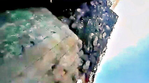 Blurry pixelated green foam crashing on purple rocks with blue sky in adult video frame
