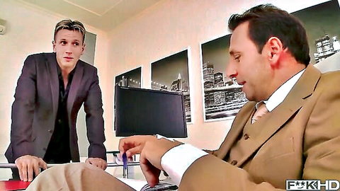 Two suited businessmen in office with younger guy leaning over older at desk teasing foreplay