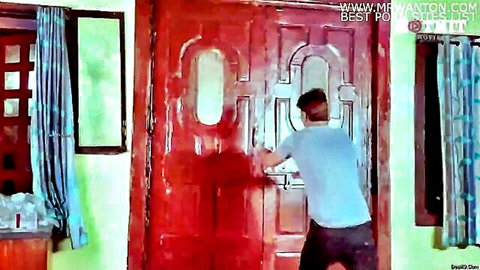 South Asian man in blue shirt and jeans forcefully pushing large red wooden door in green room