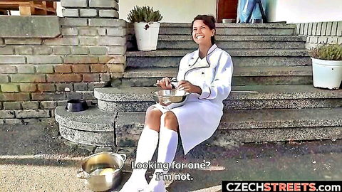 Brunette nurse babe in short white dress and stockings sits smiling on outdoor steps holding cooking pan teasing