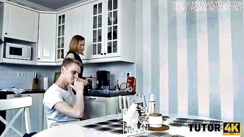 Blonde tutor in black dress stands behind young male student in kitchen during lesson