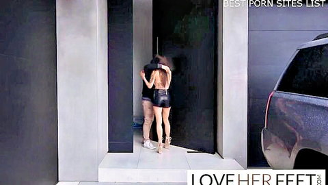 Maddy May in sexy black minidress and heels, embraced from behind in foot fetish tease