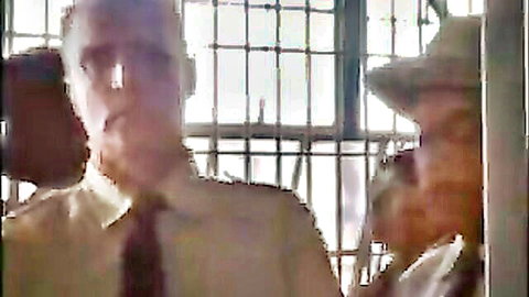 Bearded mature man in shirt and tie talks to older hatted man through prison bars