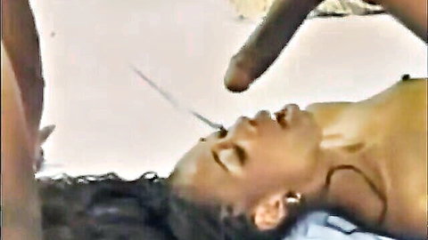 Ebony woman lies back with massive black cock hovering over her open mouth for deep throat face fuck