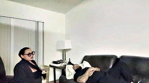 Betty Bang in glasses and cleavage-giving blazer, advising shirtless man on couch